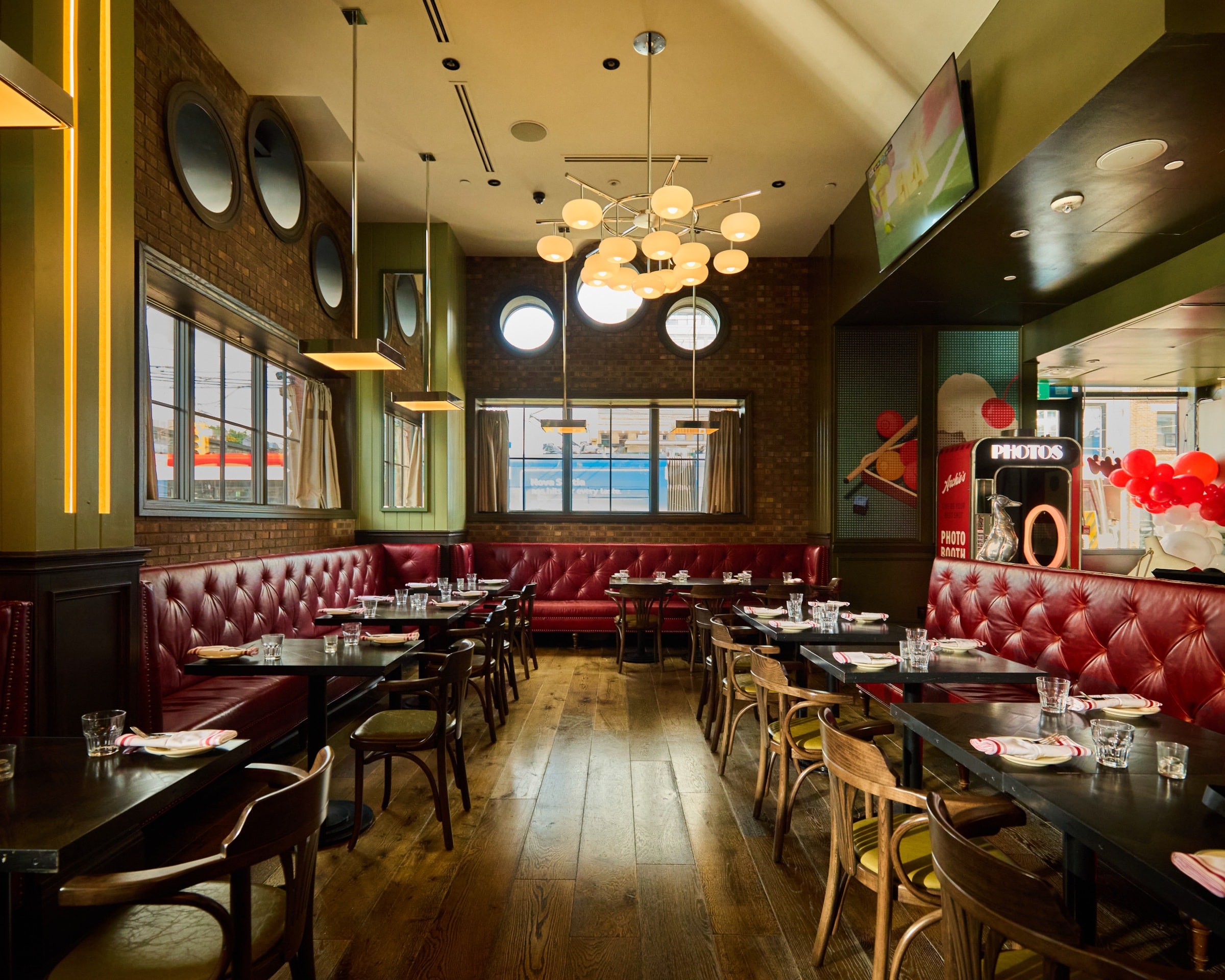 Wide view of Archie's dining room with set tables, red leather banquettes, wood floors, globe chandeliers, and the photo booth
