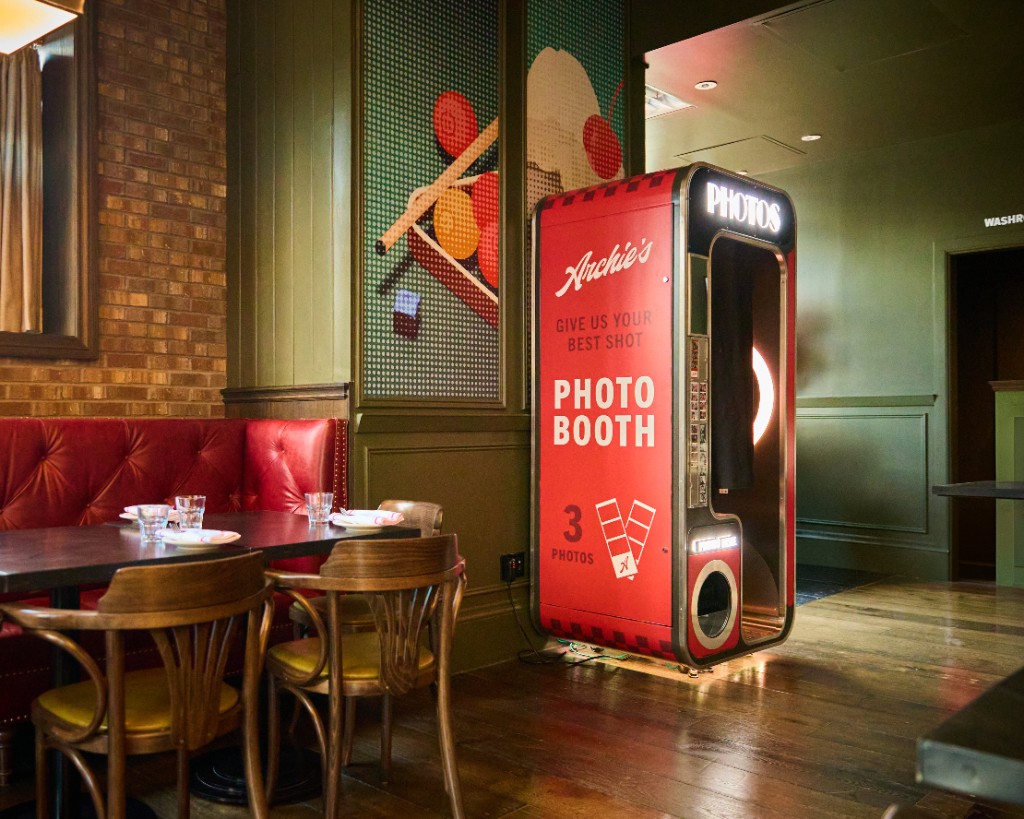 Red Archie's photo booth beside dining tables, red banquettes, brick, and green panelled walls