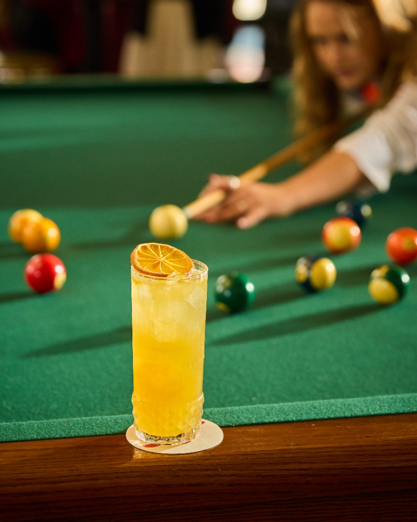 Yellow citrus cocktail on the rail of a green pool table while a guest lines up a shot